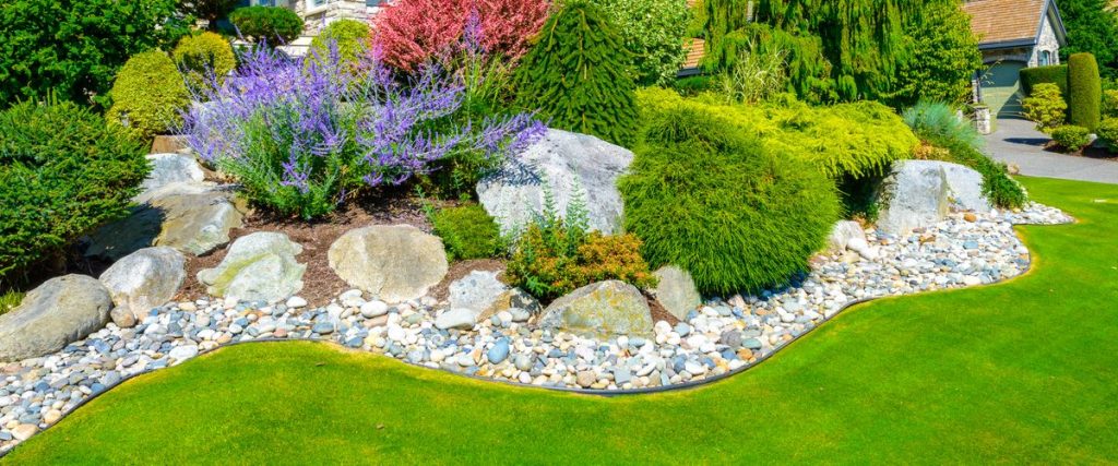 Close-up of decorative, interlocking stone pavers used as a border between a lush green lawn and a flowerbed containing colorful plants.