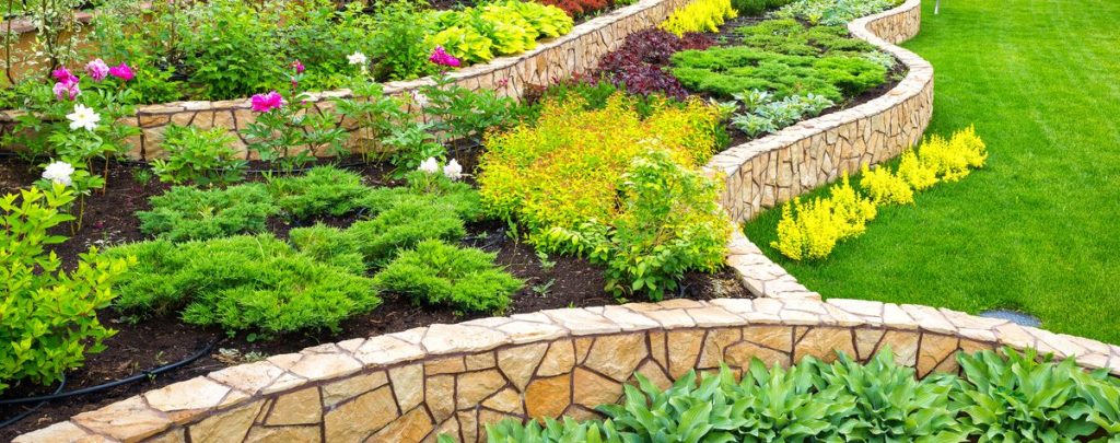 A multi-tiered flower bed design featuring winding, stacked stone retaining walls used for edging, separating colorful shrubs and flowers from a bright green lawn.