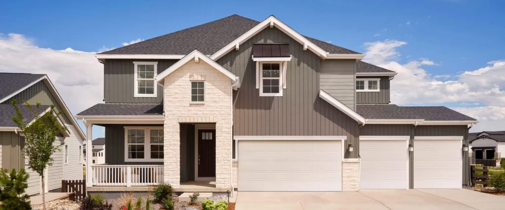 Exterior view of a modern two-story craftsman home at Macanta by Taylor Morrison featuring grey vertical siding, white stone accents, a front porch with white railings, and a spacious three-car garage under a clear blue sky.
