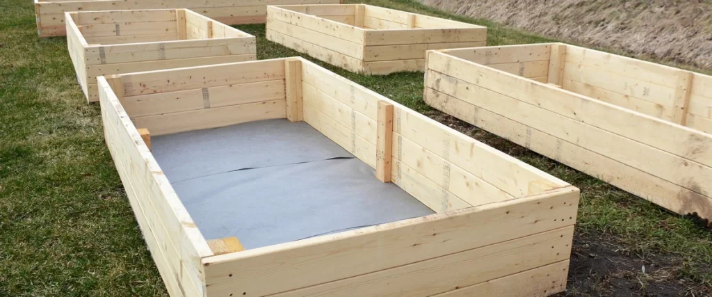 Several newly constructed, empty wooden raised garden bed frames sitting on a lawn, with one bed in the foreground showing a grey weed barrier fabric lining the bottom.