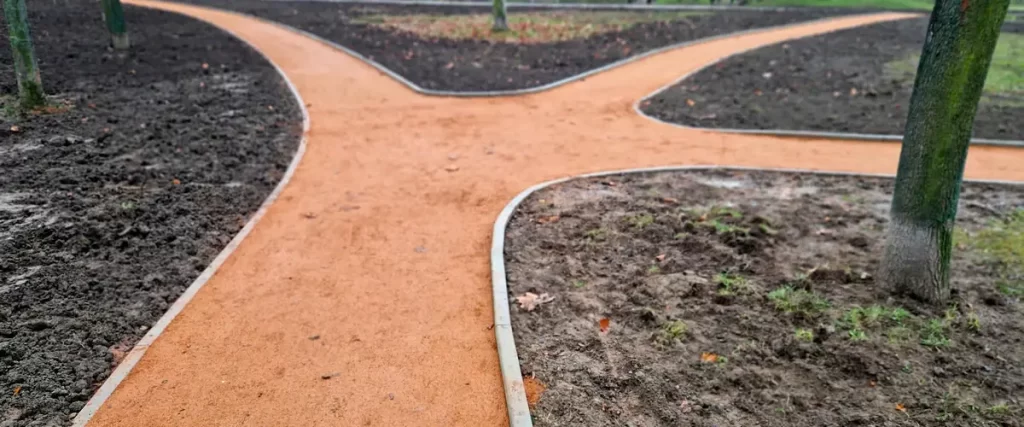 Intersecting garden paths with metal edging filled with smooth, compacted orange decomposed granite surrounded by bare soil and trees.