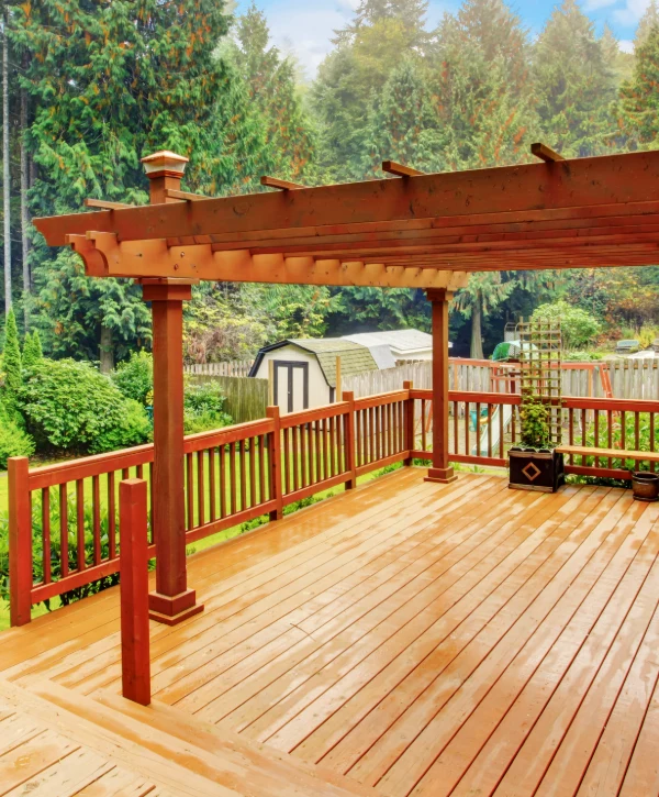 A spacious wet wooden deck featuring a classic reddish-brown wood pergola and matching railings, built by a custom pergola builder in Centennial, CO, surrounded by tall green pine trees.