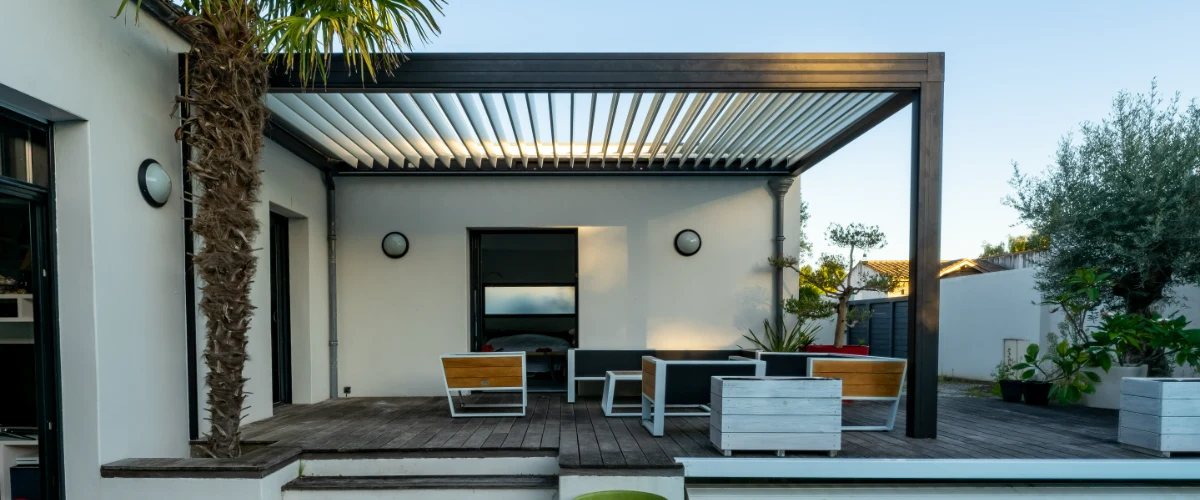 A modern dark metal pergola with a louvered roof attached to a white stucco house, shading a wooden deck with contemporary outdoor lounge furniture and a palm tree.